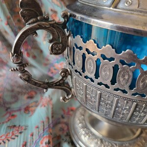 Peut inclure: Un bol antique argenté avec un intérieur en verre bleu vif. Le bol présente des détails de filigrane complexes, une poignée décorative avec un motif de papillon et un couvercle assorti. Le design orné ajoute une touche d'élégance.