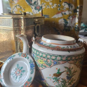 Puede incluir: Un juego de té de cerámica blanca con tapa y platillo pequeño. El juego está decorado con motivos florales y de pájaros coloridos. Una caja de metal dorada está en el fondo. El juego de té está sobre una superficie marrón.