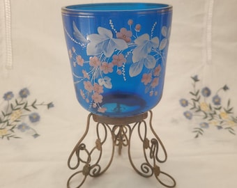 May include: A vibrant blue glass vase with floral designs in white and pink, set on an ornate, antique-style metal stand. The vase has a cylindrical shape and a gold rim. The stand has a decorative, scrolled design.