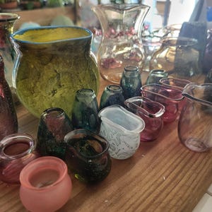 Peut inclure: Une collection de vases et de petits récipients en verre de différentes formes et tailles, dans des tons de vert, de rose et transparent. Certains ont des surfaces texturées. L'ensemble est disposé sur une surface en bois.