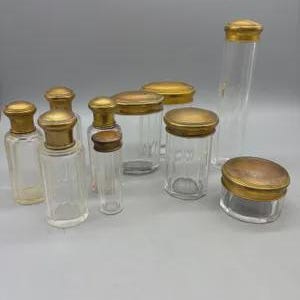 May include: A set of nine vintage glass jars with gold lids. The jars are clear glass and have a variety of shapes and sizes. The lids are all gold metal and have a simple design.