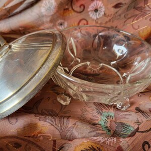 Op de afbeelding: Een Art Deco stijl glazen en metalen snuisterijenschaal. De heldere glazen kom staat op drie kleine voetjes. Het deksel is zilverkleurig metaal met een geometrisch ontwerp. De schaal is open en onthult de binnenkant.