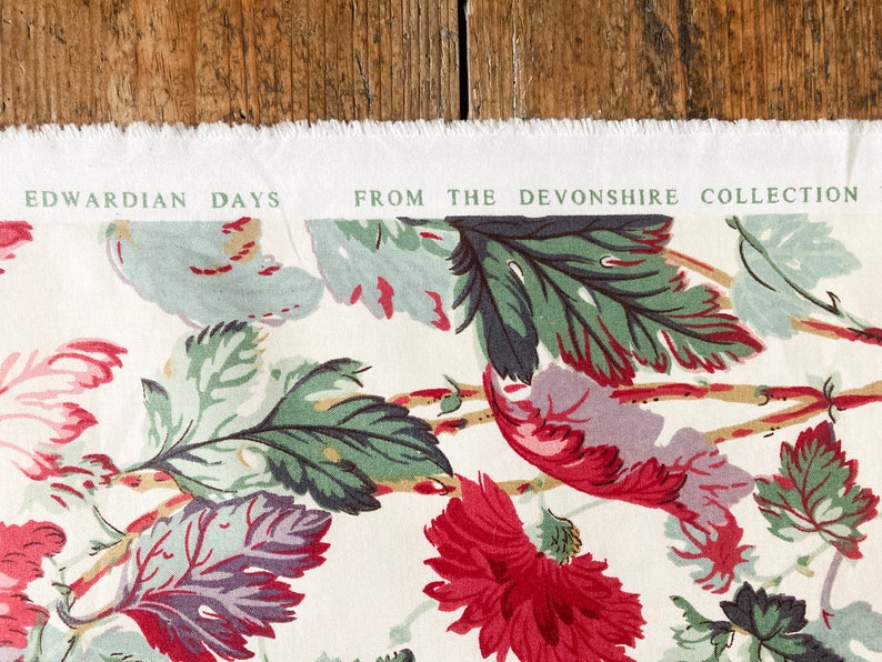 May include: A white fabric with a floral pattern featuring green, red, and purple leaves. The fabric has a label that reads "Edwardian Days from the Devonshire Collection."