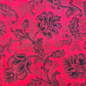 以下が含まれることがあります： 赤色の生地のクローズアップ。黒色の花柄が繰り返されています。 大きく様式化された花と葉が特徴で、複雑なディテールがあり、ヴィンテージな美しさを醸し出しています。 生地は質感のある外観をしています。