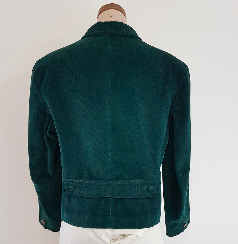 K&ouml;nnte beinhalten: Ein dunkelgr&uuml;ner Cordblazer mit einem einzigen Knopfverschluss und zwei Taschen auf der Vorderseite. Der Blazer hat eine klassische Passform und ist aus einem weichen, bequemen Stoff gefertigt.