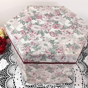 Peut inclure: Une boîte de rangement hexagonale avec un motif floral dans les tons de rose, vert et blanc. La boîte a une bordure bordeaux et un motif floral assorti sur le couvercle et les côtés. Posée sur une nappe en dentelle blanche.