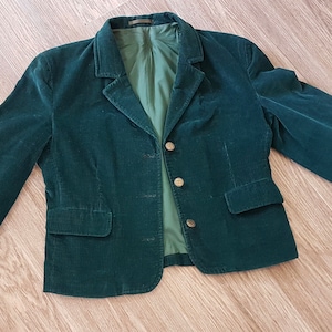 K&ouml;nnte beinhalten: Ein dunkelgr&uuml;ner Cordblazer mit einreihigem Verschluss und drei goldenen Kn&ouml;pfen. Der Blazer hat zwei Eingrifftaschen und ein Futter.
