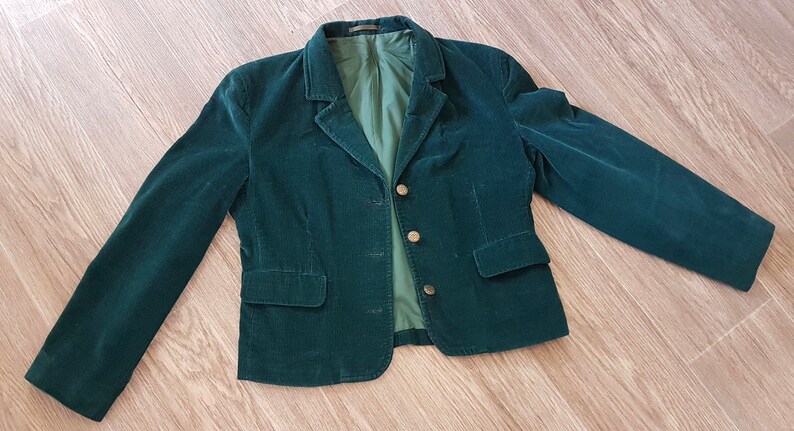 K&ouml;nnte beinhalten: Ein dunkelgr&uuml;ner Cordblazer mit einreihigem Verschluss und drei goldenen Kn&ouml;pfen. Der Blazer hat zwei Eingrifftaschen und ein Futter.
