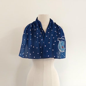 May include: A navy blue scarf with white polka dots and a decorative patch. The patch features a blue vase with a red flower and green leaves. The scarf is draped over a mannequin.