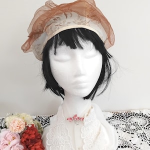 May include: A white and brown patterned hat with a sheer brown veil. The hat is on a mannequin with black hair.