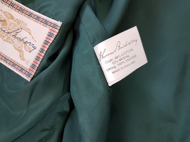 K&ouml;nnte beinhalten: Wei&szlig;es Etikett mit schwarzem Text, der "Thomas Burberry" lautet und die Stoffzusammensetzung detailliert: "Cloth: 88% Baumwolle 12% Modal" und "Lining: 100% Viskose". Das Etikett tr&auml;gt au&szlig;erdem den Hinweis "Made in England".