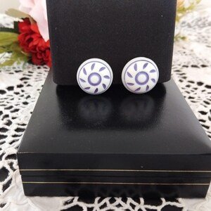 May include: A pair of round, white button-style earrings featuring a central purple circle surrounded by a ring of small, curved purple petal-like shapes. The earrings have a smooth, matte finish and are displayed on a black rectangular stand.
