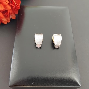 May include: A pair of silver-coloured clip-on earrings. Each earring features a textured, curved design with a small, round accent at the base. The earrings are presented on a black surface, with a hint of orange flowers in the background.
