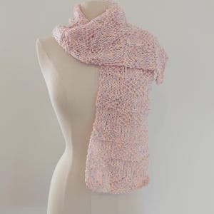 May include: A pale pink knitted scarf on a mannequin. The scarf features a textured design and is made from a soft yarn with flecks of blue, yellow, and pink. The scarf is draped to show its length and pattern.