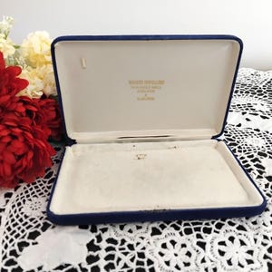 May include: A rectangular, hinged jewellery box with a deep blue exterior and a cream-coloured interior lining. The inside lid features gold text reading WENDY'S JEWELLERS, 74 RUNDLE MALL, ADELAIDE & SUBURBS. The box is empty and has a soft, velvet-like texture.