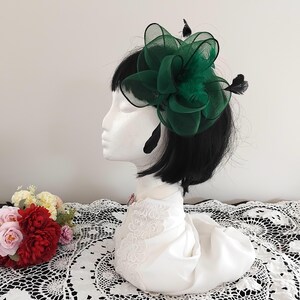 May include: A striking dark green fascinator, featuring a large, layered floral design and black feathers. The headpiece is displayed on a white mannequin head with black hair. This accessory is ideal for formal events and adds a touch of sophistication.