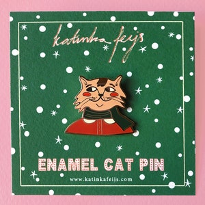 K&ouml;nnte beinhalten: Eine gr&uuml;ne Karte mit wei&szlig;en Punkten und goldenen Sternen zeigt eine rote und gr&uuml;ne Emaille-Anstecknadel mit einer Katze, die einen roten Pullover und einen gr&uuml;nen Schal tr&auml;gt. Der Text "katinha feijs" und "ENAMEL CAT PIN" ist in Gold auf der Karte gedruckt. Die Website-Adresse "www.katinkafeijs.com" ist in Gold am unteren Rand der Karte gedruckt.