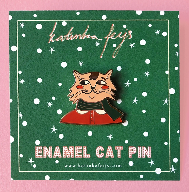 K&ouml;nnte beinhalten: Eine gr&uuml;ne Karte mit wei&szlig;en Punkten und goldenen Sternen zeigt eine rote und gr&uuml;ne Emaille-Anstecknadel mit einer Katze, die einen roten Pullover und einen gr&uuml;nen Schal tr&auml;gt. Der Text "katinha feijs" und "ENAMEL CAT PIN" ist in Gold auf der Karte gedruckt. Die Website-Adresse "www.katinkafeijs.com" ist in Gold am unteren Rand der Karte gedruckt.