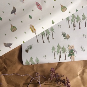 Op de afbeelding: Twee vellen wit inpakpapier met verschillende ontwerpen. Het bovenste vel heeft een herhalend patroon van vogels, huizen en kerstboompjes. Het onderste vel heeft een herhalend patroon van dennenbomen, een kat die een dennenboom vasthoudt, en dennenappels.