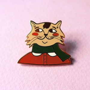 K&ouml;nnte beinhalten: Eine goldfarbene Emaille-Anstecknadel mit einem Cartoon-Katzenmotiv, das einen roten Pullover und einen gr&uuml;nen Schal tr&auml;gt. Die Katze hat braunes Fell, einen wei&szlig;en Kragen und einen freundlichen Gesichtsausdruck.