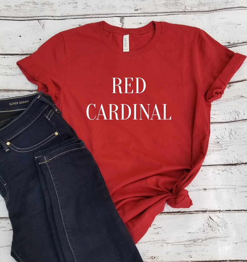 Puede incluir: Camiseta roja con el texto "RED CARDINAL" en letras blancas. La camiseta se combina con unos vaqueros azul oscuro.