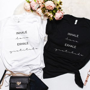 May include: Two t-shirts with the text "INHALE love EXHALE gratitude" printed on them. One t-shirt is white and the other is black.