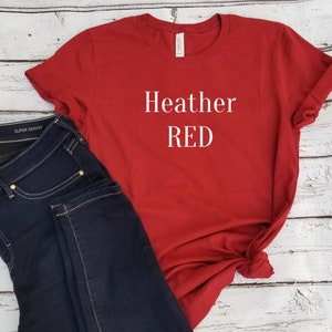 May include: Red t-shirt with the text "Heather RED" printed in white on the front. The shirt is paired with dark blue jeans.