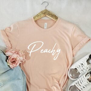 May include: Peachy in white script on a light pink t-shirt. The t-shirt is hanging on a gold hanger.