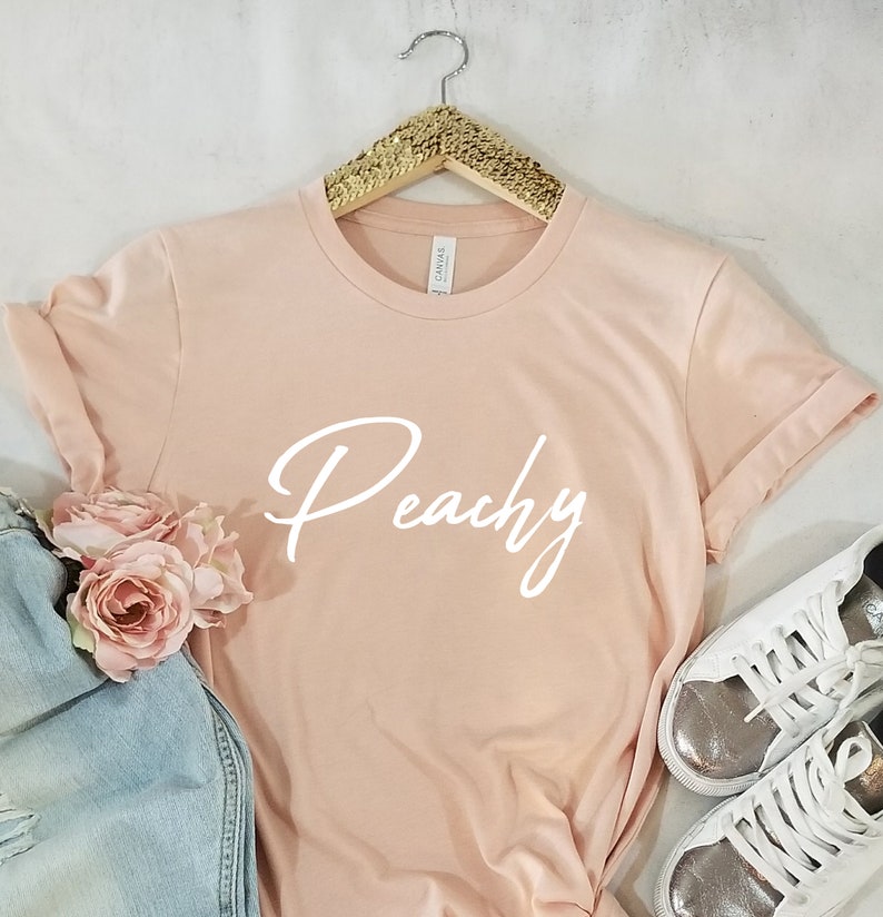 May include: Peachy in white script on a light pink t-shirt. The t-shirt is hanging on a gold hanger.