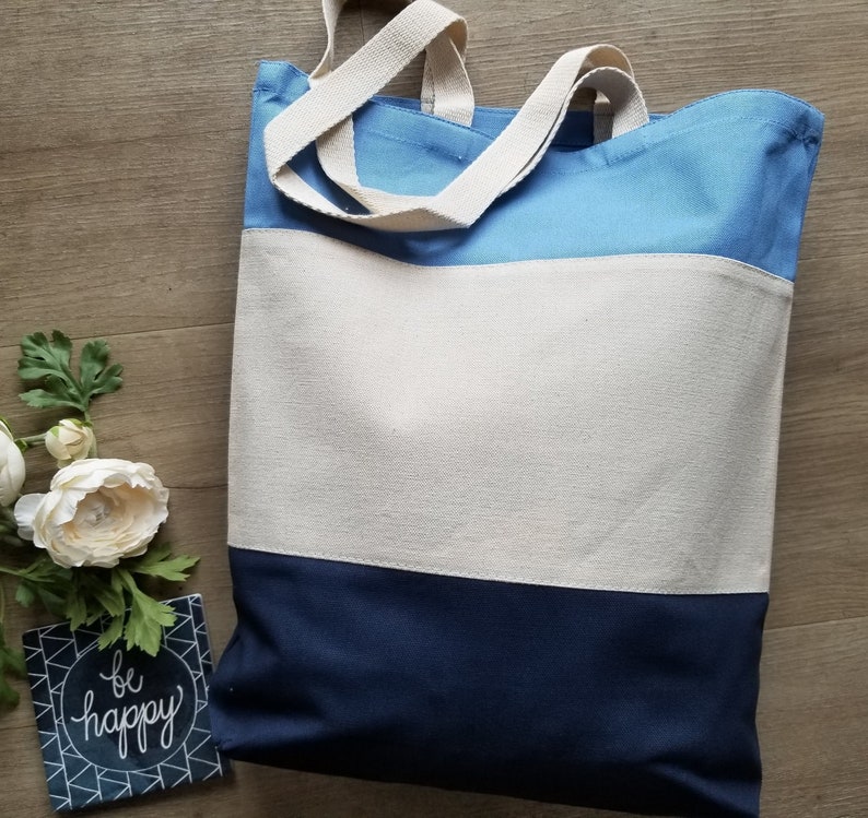 May include: A blue, white, and navy blue canvas tote bag with two long handles. The bag has the text "be happy" printed on a small black and white tile in the foreground.