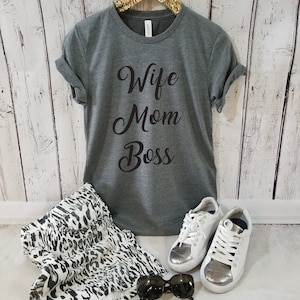 Puede incluir: Una camiseta gris con el texto "Wife Mom Boss" en letras cursivas negras. La camiseta est&aacute; en una percha y hay zapatillas blancas, gafas de sol y una falda de estampado animal blanco y negro en la imagen.