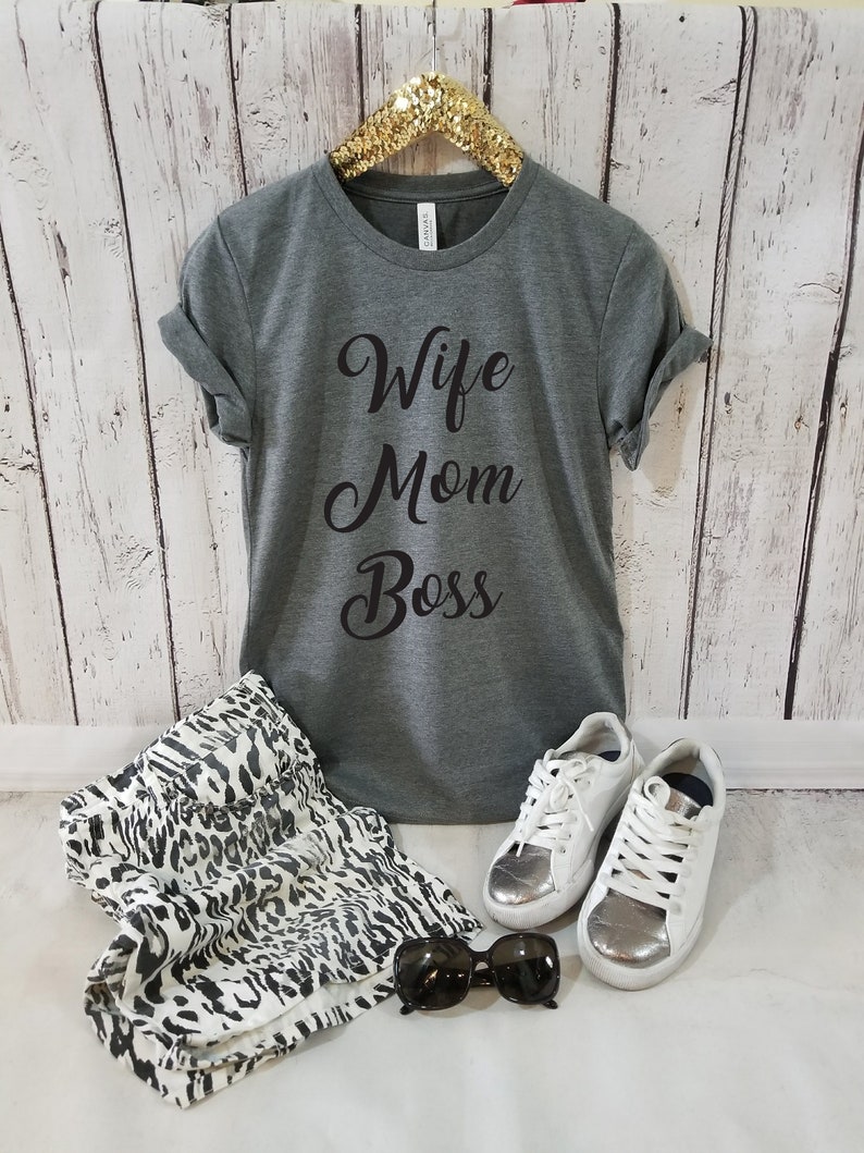 Puede incluir: Una camiseta gris con el texto "Wife Mom Boss" en letras cursivas negras. La camiseta est&aacute; en una percha y hay zapatillas blancas, gafas de sol y una falda de estampado animal blanco y negro en la imagen.