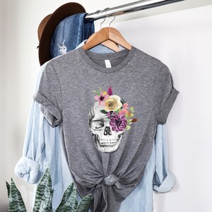 May include: Gray t-shirt with a floral skull design. The skull is white with black eye sockets and a black nose. The flowers are pink, yellow, purple, and green.
