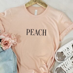 May include: Peach colored t-shirt with the word "PEACH" printed in black on the front. The shirt is laying on a white surface with a pair of white sneakers with silver accents.