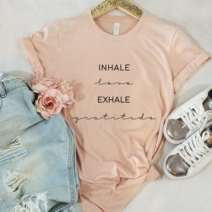 May include: A pink t-shirt with the text "INHALE love EXHALE gratitude" printed on it. The shirt is on a white background with a pair of jeans and white sneakers.