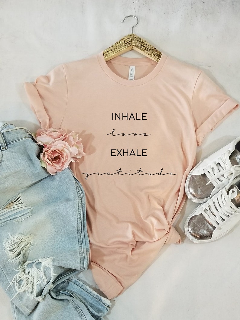 May include: A pink t-shirt with the text "INHALE love EXHALE gratitude" printed on it. The shirt is on a white background with a pair of jeans and white sneakers.