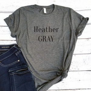 May include: Heather Gray is printed in black on a heather gray t-shirt. The t-shirt is folded and laid on a white wooden surface. A pair of blue jeans is partially visible in the bottom left corner of the image.