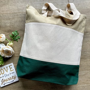 May include: A beige and green canvas tote bag with two handles. The bag has a large white section in the middle with the words "LOVE the JOURNEY" printed on a small white square in the bottom right corner.