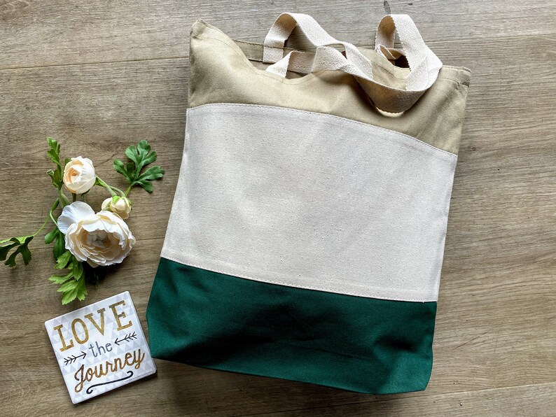 May include: A beige and green canvas tote bag with two handles. The bag has a large white section in the middle with the words "LOVE the JOURNEY" printed on a small white square in the bottom right corner.