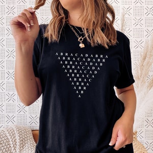 May include: Black t-shirt with a white graphic design of the word "ABRACADABRA" repeated in a pyramid shape.