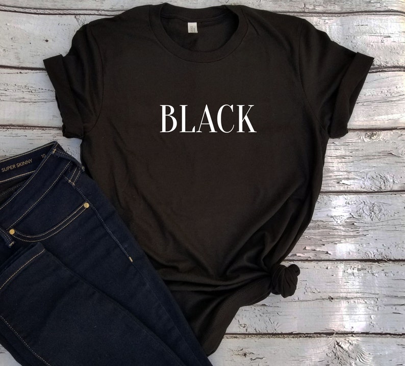 May include: Black crew neck t-shirt with the word "BLACK" in white block letters. Dark blue skinny jeans are next to the shirt. The shirt is folded at the sleeves.