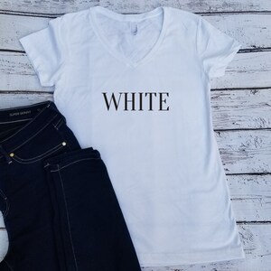 May include: White v-neck t-shirt with the word "WHITE" printed in black on the front. The shirt is laying on a wooden surface with a pair of dark blue jeans.