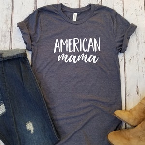 May include: A dark gray t-shirt with the words "AMERICAN mama" in white script. The shirt is folded and placed on a light wood-grain surface, next to a pair of blue jeans and tan boots.