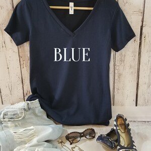 May include: A navy blue v-neck t-shirt with the word "BLUE" printed in white on the front. The shirt is hanging on a gold hanger and is displayed on a white wood background.