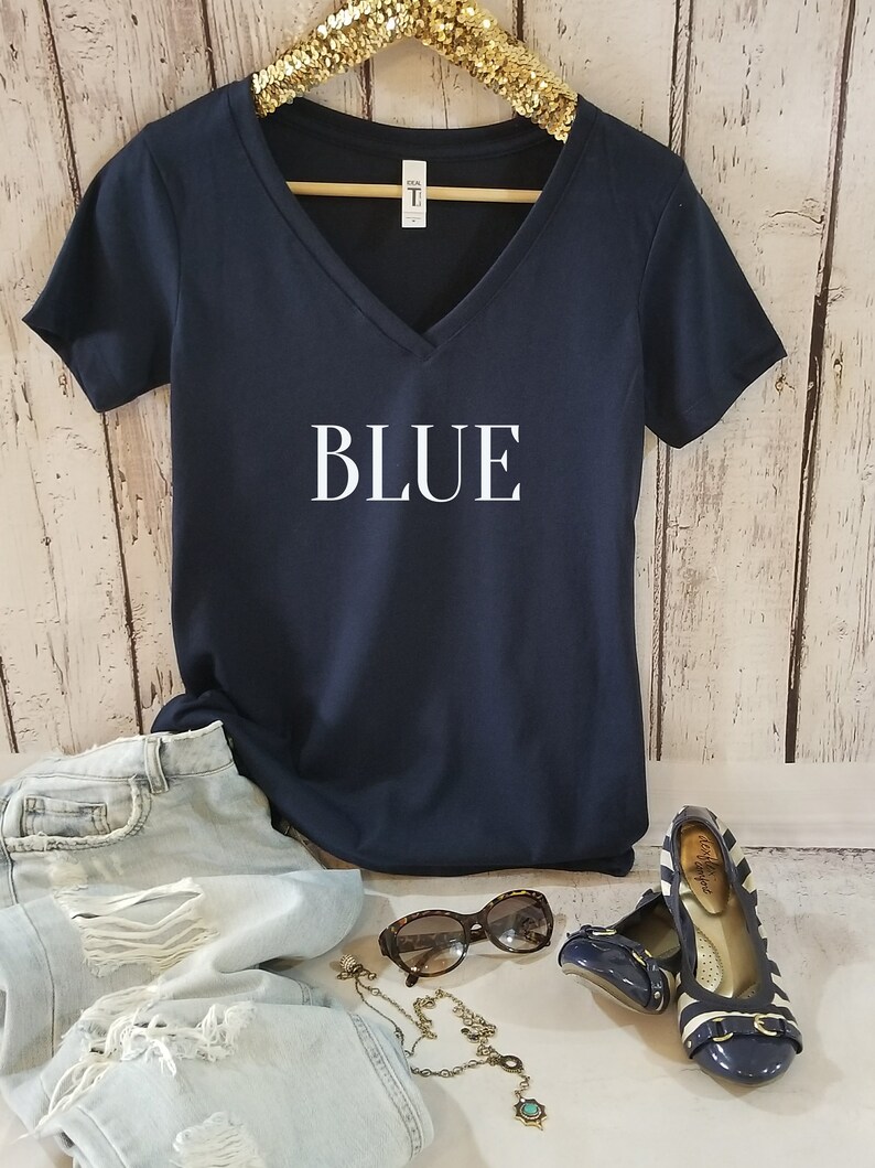 May include: A navy blue v-neck t-shirt with the word "BLUE" printed in white on the front. The shirt is hanging on a gold hanger and is displayed on a white wood background.