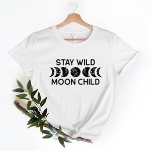 Peut inclure: T-shirt blanc avec un texte noir qui dit "Stay Wild Moon Child" et un graphique de cinq phases de la lune.