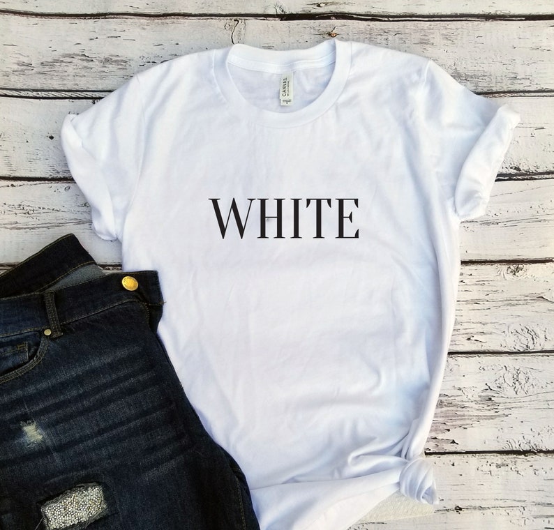 以下が含まれることがあります： 白いTシャツに、黒い文字で「WHITE」とプリントされています。Tシャツは白い木製の表面に置かれ、左側に青いジーンズがあります。