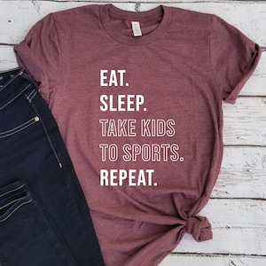 May include: A maroon t-shirt with white text that reads "Eat. Sleep. Take Kids To Sports. Repeat."