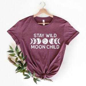 Peut inclure: Un t-shirt marron avec le texte "Stay Wild Moon Child" en lettres blanches. Le texte est accompagn&eacute; d'un graphique d'une lune dans diff&eacute;rentes phases.