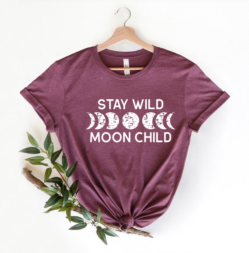 Peut inclure: Un t-shirt marron avec le texte "Stay Wild Moon Child" en lettres blanches. Le texte est accompagn&eacute; d'un graphique d'une lune dans diff&eacute;rentes phases.